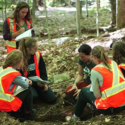 Six étudiantes portant un dossard qui sont accroupies en forêt et lisent des notes.